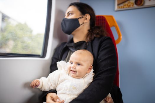 Woman wearing a face mask and black clothes and looking through the window of a train as she holds her smiling baby