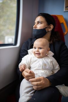 Woman wearing a face mask and black clothes and looking through the window of a bus as she holds her smiling baby