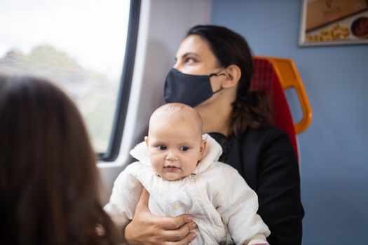 Woman wearing a face mask and looking through the window of a bus as she holds her smiling baby in her arms