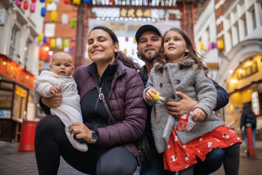 Picture of a smiling woman holding her baby daughter alongside her smiling husband who hugs his daughter, with a blurry Chinese gate from Chinatown as background