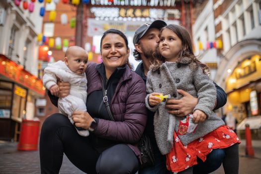 Picture of a smiling woman holding her baby daughter alongside her husband who hugs and kisses his daughter, with a blurry Chinese gate from Chinatown as background