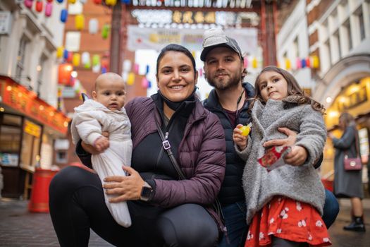 Picture of a smiling woman holding her baby daughter and posing alongside her smiling husband who hugs his daughter, with a blurry Chinese gate from Chinatown as background