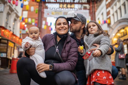 Picture of a smiling woman holding her baby daughter alongside her husband who kisses her while he hugs his daughter, with a blurry Chinese gate from Chinatown as background