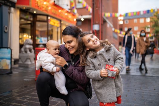 Picture of a smiling little girl resting her head on the shoulder of her mother who is kneeling and holding her baby daughter, with a blurry alley from Chinatown and people walking as the background