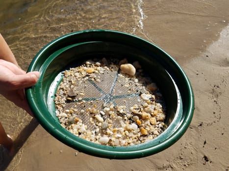 female hand holding strainer with stones at beach with water and sand