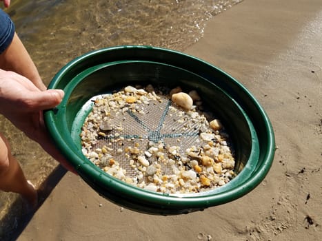 female hand holding strainer with stones at beach with water and sand