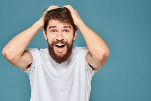 A bearded man in a white T-shirt gestures with his hands emotions blue background. High quality photo