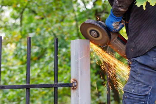 A worker cuts a metal post with a disc angle grinder and sets a fence against a blurred green park, creating a bright plume of many hot sparks.