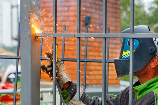 A welder wearing a safety helmet and gloves is welding a metal fence around a residential building, bright sparks fly, blue smoke, selective focus, copy space.