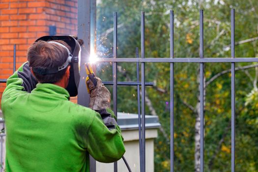 A welder in a protective helmet and gloves using an electrode welds a metal fence in a park area around a residential building, bright sparks, blue smoke fly, selective focus, copy space.