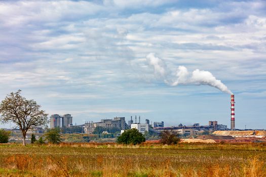 Cement plant on the horizon with a smoking chimney and a quarry amid agricultural field and blue cloudy sky, copy space.
