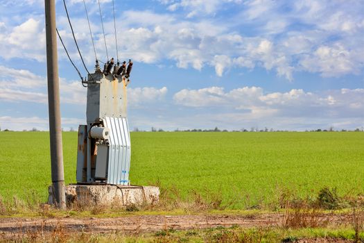 An old transformer station stands in the middle of a green field, near a concrete pillar, against a blue cloudy sky.