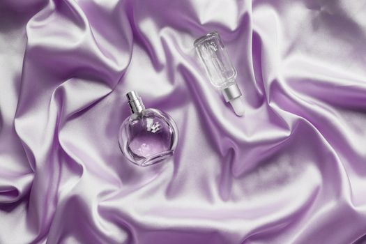 Perfume bottle and face serum on lilac silk folded fabric background. Luxery Scent fragrance cosmetic beauty product.