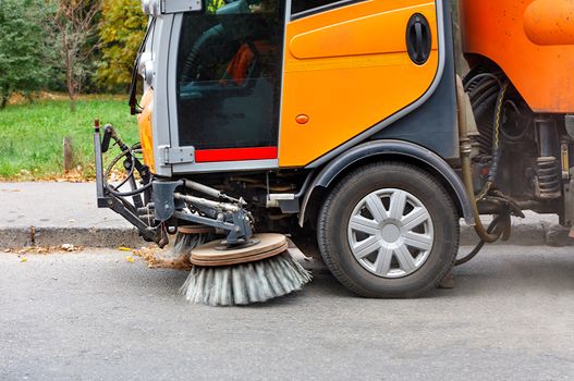 Sweeper's rotating front brushes pick up debris and fallen leaves from the carriageway and near the curb, copy space.