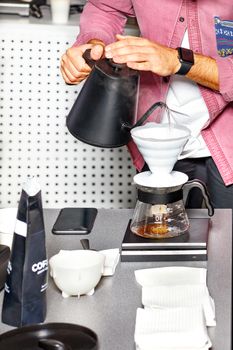 Male hands of barista prepare coffee by pouring boiling water from a black kettle into a white funnel of a glass flask.