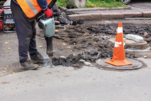 A road worker, dressed in reflective clothing, uses an electric jackhammer to rip old asphalt off the road and uses a gasoline generator, copy space.