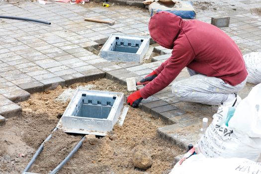 A worker installs engineering hatch shafts and lays paving slabs on prepared flat sandy soil on the sidewalk, copy space.