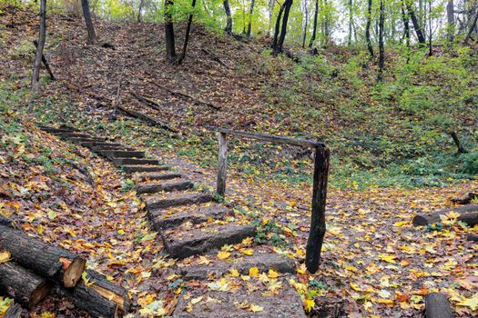 Autumn forest. An old stone staircase with a fragment of a wobbly wooden railing strewn with leaves turns and climbs up.