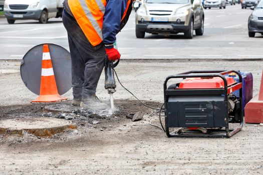 A road worker, dressed in reflective clothing, repairs a section of road near sewers with an electric jackhammer, with passing cars in blur as background, copy space.