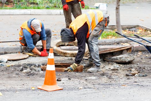 Road workers on a fenced-in stretch of road, dressed in reflective clothing, replace an old sewerage manhole.