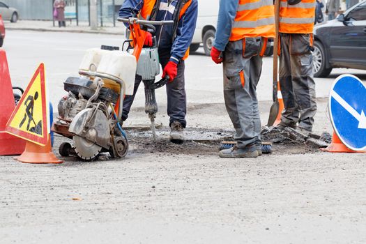 Road workers in reflective clothing on a fenced-in section of the road repair the roadway with an electric jackhammer, petrol cutter, shovel and brush. Copy space.