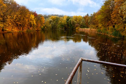 The rusted metal railings of the old bridge direct the gaze to a beautiful autumn forest lake landscape with waterfowl wild ducks in blur.