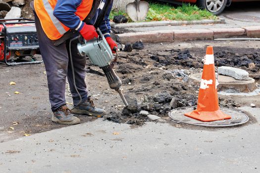 A road worker in reflective clothing breaks old asphalt off the road with an electric jackhammer.