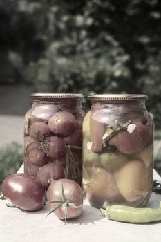 Home preservation: two large glass jars with red ripe pickled tomatoes and peppers, closed with a metal lid, on a table in the garden. Front view, copy space