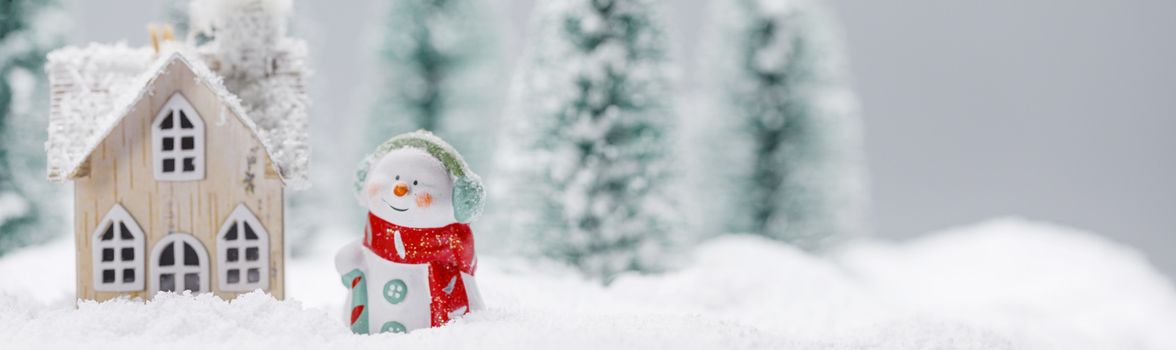 Small decorative snowman near wooden house in fir forest under falling snow