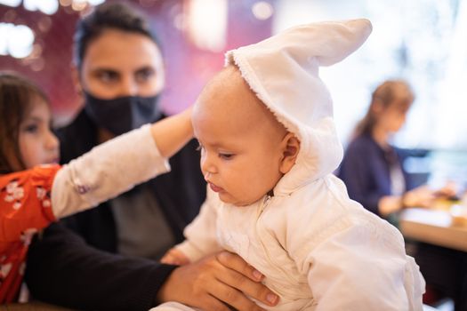 Baby in white clothing sitting on a cafe table alongside her mother and older sister