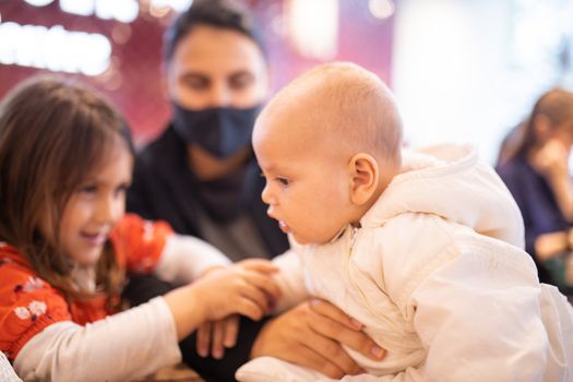 Picture of a happy little brunette girl holding the arm of her baby sibling who is wearing white clothing and sitting in profile on a table of a Chinese restaurant, and with their mother behind them