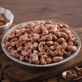 Cashew nuts with peel in a plate on wooden tray and table background, healthy raw food concept.