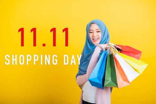 Asian Muslim Arab, Portrait of happy beautiful young woman Islam religious wear veil hijab funny smile she holding colorful shopping bags with 11.11 big sale text at side isolated on yellow background