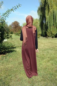Woman in a hijab. Girl in traditional muslim brown clothes on a background of green grass. Stands with your back.