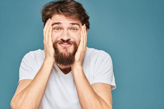 A bearded man in a white T-shirt gestures with his hands emotions blue background. High quality photo