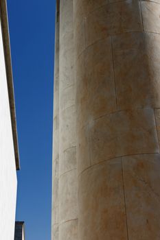 A patch of blue sky between a row of round columns in backlight and a white wall in full sun , vertical composition , wide angle lens from the bottom to the top 