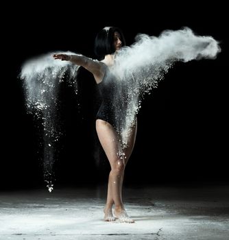woman with athletic figure dancing in flying white flour on black background, the dancer is wearing a black bodysuit