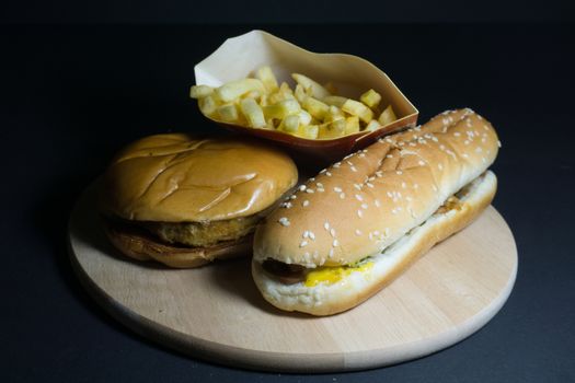 French fries with hamburger and hot dog on wooden tray with black background