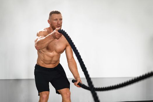 Gym battle rope man stamina training Athlete guy fitness exercising endurance indoor workout. Handsome caucasian sportive guy doing exercise functional training fitness. Muscular men naked torso