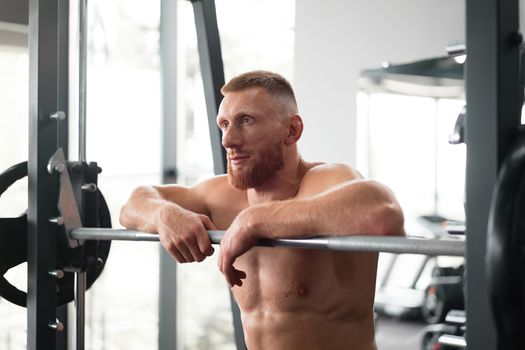 Gym man leaning barbell resting after hard training. Handsome caucasian athlete take break. Sport equipment Sportive guy standing near barbell machine. Perfect muscule male body ideal. Naked torso