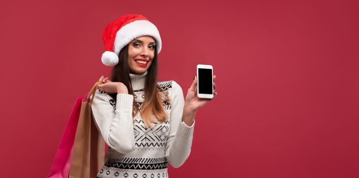 Portrait beautifiul caucasian woman Santa hat on red studio background. Christmas New Year holiday concept. Cute girl teeth smiling positive emotions with shopping bag and smartphone Online shopping