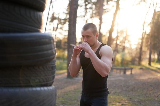 Man fighter training boxing outdoor tire DIY handmade gym in forest fitness workout. Young adult training right hand hit nature without boxing gloves. Sportive activie male healthy lifestyle concept.