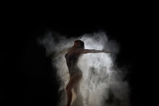 beautiful young caucasian woman in a black bodysuit with a sports figure is dancing in a white cloud of flour on a black background, explosion and expresse in motion