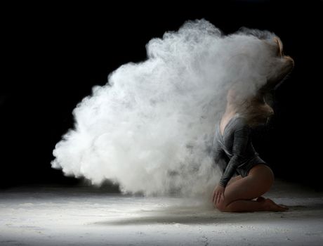 beautiful girl in a black bodysuit with long hair sits and raises her head up, white flour flies in different directions on a black background, explosion