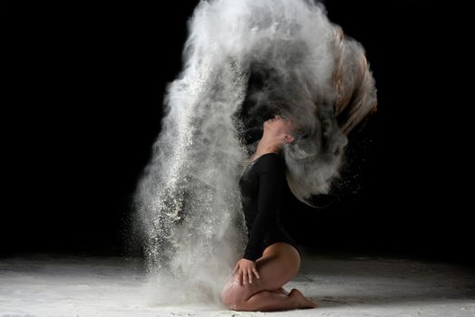 young beautiful woman with long hair is dressed in a sports black bodysuit and sits on the floor and throwing white flour up, black background