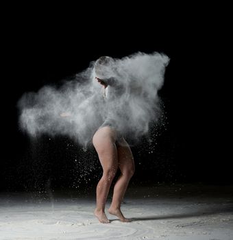 beautiful young caucasian woman in a black bodysuit with a sports figure is dancing in a white cloud of flour on a black background
