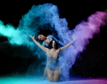 two beautiful young caucasian women in black bodysuits with a sports figure are dancing in a purple-blue cloud of flour on a black background, explosion and expresse in motion