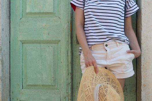 Fashinable torso portrait of woman holding sun hat, standing and relaxing in shade in front of turquoise vinatage wooden door in old Mediterranean town while sightseeing on hot summer day.