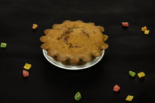 On a dark surface there is a white saucer with a chocolate cake and scattered colorful candied fruits