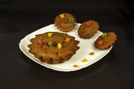 On a dark background there is a white square plate with chocolate cakes and candied fruits